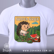 Футболка їжачок оптиміст. Біла футболка, складена на поверхні, з принтом їжачка, що тримає кавун. На фоні видно зелені пагорби та блакитне небо, принт яскравий і детальний, з елементами природи.