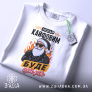 Футболка кайфовий новий рік, біла, зображена зверху, фон світло-сірий, принт з дідусем у чорній куртці, текст "НЕХАЙ КАЙФОВИМ БУДЕ ЦЕЙ РІК", на футболці є етикетка з зеленим шнурком.