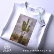 Футболка кактус обійми, біла, зображена зверху, на фоні світло-сірого, принт з кактусом та написом 'HUG ME', на комірці є етикетка.