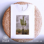 Футболка кактус обійми, біла, складена на плетеному кошику, зображена зверху, фон світло-сірий, принт з кактусом та написом 'HUG ME'.