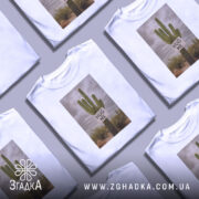 Футболка кактус обійми, біла, зображена з фронтального ракурсу, на фоні сірого градієнта, принт з кактусом та написом 'HUG ME' на білому фоні.