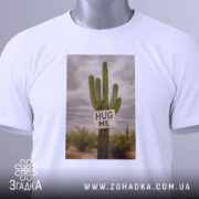 Футболка кактус обійми, біла, зображена зверху, на фоні світло-сірого, принт з кактусом та написом 'HUG ME', на комірці є етикетка.