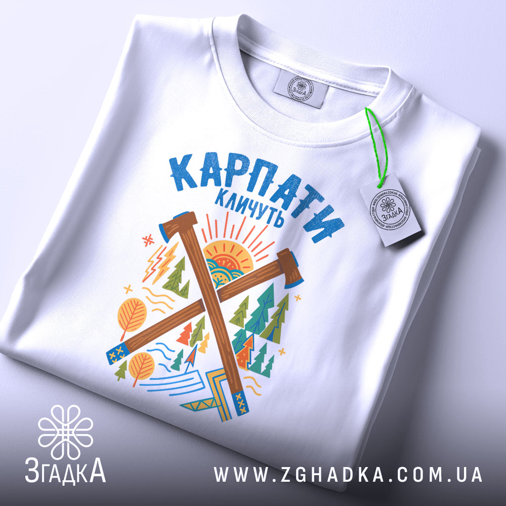 Футболка Карпати кличуть подарунок, біла футболка складена на столі, фон світло-сірий, принт з лісом та сонцем, на футболці є етикетка з зеленим шнурком.