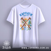 Футболка Карпати кличуть подарунок, біла футболка підвішена на вішалці, фон світло-сірий, принт з лісом та сонцем, яскраві кольори принта.