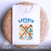 Футболка Карпати кличуть подарунок, біла футболка складена на плетеній кошику, фон світло-сірий, принт з лісом та сонцем, яскраві кольори принта.