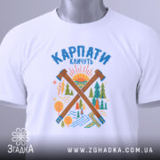 Футболка Карпати кличуть подарунок, біла футболка складена на столі, фон світло-сірий, принт з лісом та сонцем, на футболці є етикетка з зеленим шнурком.