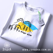 Футболка Карпати кличуть, біла, складена на поверхні, фон світлий, принт з горами, деревами та текстом, з етикеткою на комірці.