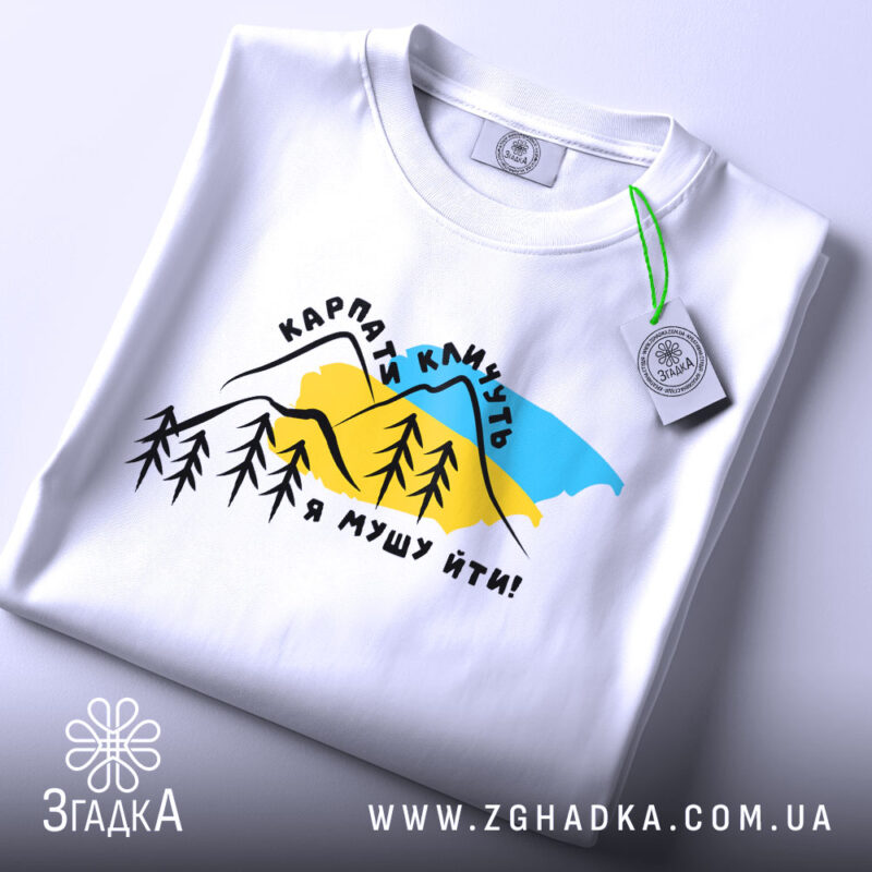 Футболка Карпати кличуть, біла, складена на поверхні, фон світлий, принт з горами, деревами та текстом, з етикеткою на комірці.