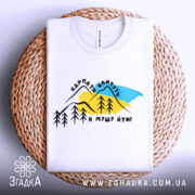 Футболка Карпати кличуть, біла, складена на плетеній основі, фон світлий, принт з горами, деревами та текстом, виглядає акуратно.