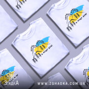 Футболка Карпати кличуть, біла, зображена на манекені, фон нейтральний, принт з горами, деревами та текстом на українській мові.