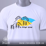 Футболка Карпати кличуть, біла, складена на поверхні, фон світлий, принт з горами, деревами та текстом, з етикеткою на комірці.
