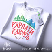 Футболка карпати кличуть, білого кольору, складена на поверхні, фон світло-сірий, принт з горами, лісом та написом червоного кольору, на комірці видна етикетка.