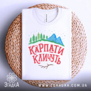 Футболка карпати кличуть, білого кольору, складена на плетеній основі, фон білий, принт з горами, лісом та написом червоного кольору.