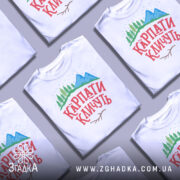 Футболка карпати кличуть, білого кольору, складена на поверхні, фон світло-сірий, принт з горами, лісом та написом червоного кольору, кілька футболок розташовані поруч одна з одною.