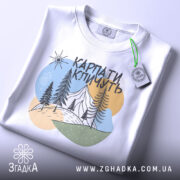 Футболка Карпати кличуть, біла, складена на поверхні, фон світло-сірий, принт з горами, лісом та сонцем, з етикеткою на зеленому шнурку.