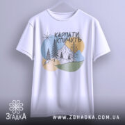 Футболка Карпати кличуть, біла, підвішена на вішалці, фон світло-сірий, принт з горами, лісом та сонцем у пастельних кольорах.
