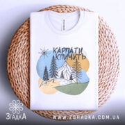 Футболка Карпати кличуть, біла, складена на плетеній кошику, фон світло-сірий, принт з горами, лісом та сонцем у пастельних кольорах.