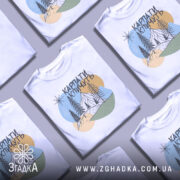 Футболка Карпати кличуть, біла, зображена на манекені, фон світло-сірий, принт з горами, лісом та сонцем у пастельних кольорах.