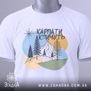 Футболка Карпати кличуть, біла, складена на поверхні, фон світло-сірий, принт з горами, лісом та сонцем, з етикеткою на зеленому шнурку.