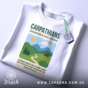 Футболка Карпати Україна, біла, складена на поверхні, фон світлий, принт з горами, сонцем і будинком, з етикеткою на рукаві.