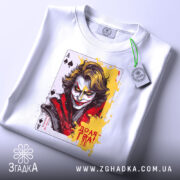 Футболка карта хаосу, біла, зображена на столі, ракурс зверху, фон світло-сірий, принт з обличчям персонажа, яскраві кольори жовтого та червоного, на комірці етикетка з логотипом.
