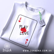 Футболка карта кохання, білого кольору, складена на столі, фон світло-сірий, принт у вигляді картки з написом 'PLAY LOVE' та червоними сердечками, на комірці є етикетка.