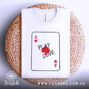 Футболка карта кохання, білого кольору, складена на плетеній кошику, фон білий, принт у вигляді картки з написом 'PLAY LOVE' та червоними сердечками.