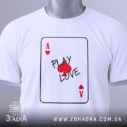 Футболка карта кохання, білого кольору, складена на столі, фон світло-сірий, принт у вигляді картки з написом 'PLAY LOVE' та червоними сердечками, на комірці є етикетка.