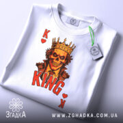 Футболка карта короля, біла, з верхнього ракурсу, на фоні світло-сірого градієнта, складена, принт з черепом у короні, червоний напис 'KING' та символи 'K' і серце, з етикеткою.