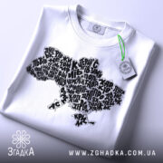 Футболка карта України білого кольору, складена на поверхні, з принтом у вигляді карти України, виконаним чорними літерами, з зеленим ярликом, на фоні світло-сірого кольору.