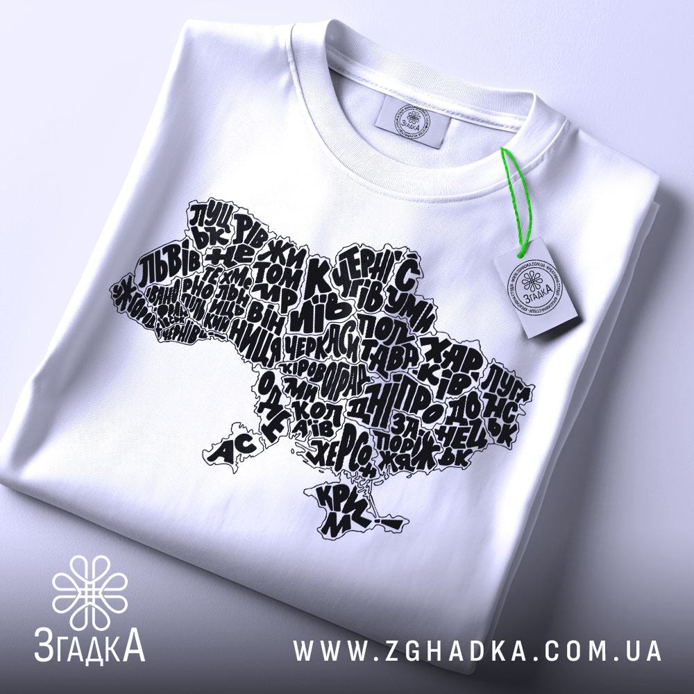 Футболка карта України білого кольору, складена на поверхні, з принтом у вигляді карти України, виконаним чорними літерами, з зеленим ярликом, на фоні світло-сірого кольору.