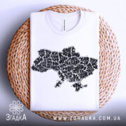 Футболка карта України білого кольору, складена на плетеній кошику, з принтом у вигляді карти України, виконаним чорними літерами, на фоні світло-сірого кольору.
