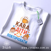 Футболка кава діти повторювати. Біла футболка з круглим вирізом, зображена зверху на білому фоні. На футболці видно принт з текстом 'Кава. Діти. Повторювати' та ілюстраціями дітей. На футболці є етикетка з зеленим шнурком.
