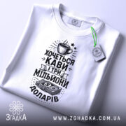 Футболка кава мільйони доларів, біла, складена на поверхні, фон світлий, принт чорного кольору з малюнком чашки кави та грошима, на комірці є етикетка.