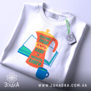 Футболка кава та книги, біла, зображена зверху, на фоні світло-сірого кольору, принт з кавоваркою, книгою та чашкою, яскраві кольори: оранжевий, жовтий, зелений, синій, текст українською мовою, з етикеткою на комірці.