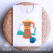 Футболка кава та книги, біла, зображена зверху на плетеній кошику, фон світлий, принт з кавоваркою, книгою та чашкою, яскраві кольори: оранжевий, жовтий, зелений, синій, текст українською мовою.