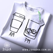 Футболка кава віскі, біла, складена на столі, фон світлий, принт з малюнком кави та склянки з льодом, підписами AM та PM, з етикеткою на рукаві.