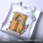 Футболка кавова магія. Біла футболка, складена на поверхні, ракурс зверху, фон світлий, принт з кавомолкою та рослинними елементами, на футболці є етикетка з зеленим шнурком.