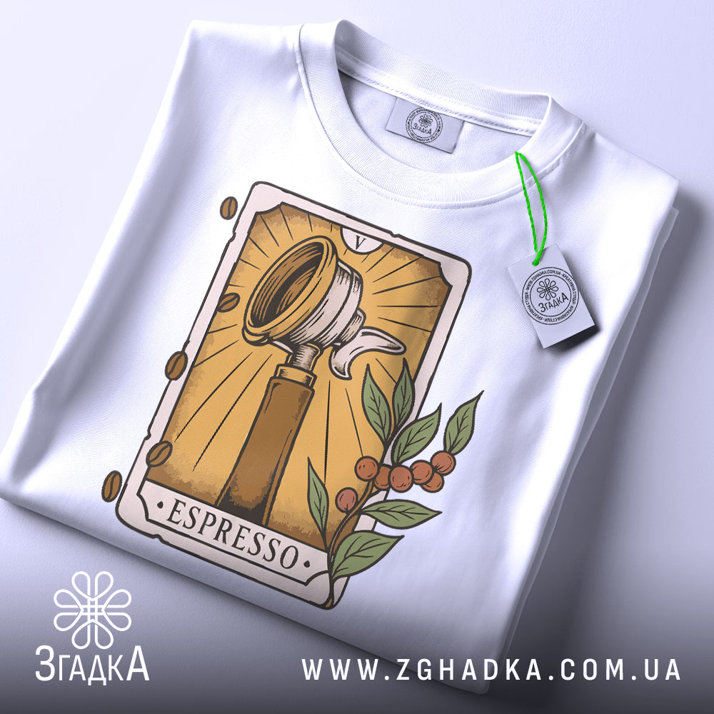 Футболка кавова магія. Біла футболка, складена на поверхні, ракурс зверху, фон світлий, принт з кавомолкою та рослинними елементами, на футболці є етикетка з зеленим шнурком.