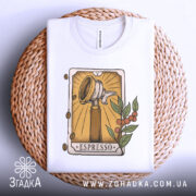 Футболка кавова магія. Біла футболка, складена на плетеній основі, ракурс зверху, фон світлий, принт з кавомолкою та рослинними елементами, видно етикетку на комірці.