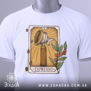 Футболка кавова магія. Біла футболка, складена на поверхні, ракурс зверху, фон світлий, принт з кавомолкою та рослинними елементами, на футболці є етикетка з зеленим шнурком.