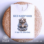 Футболка кавучіно злочіно, біла, складена на плетеному кошику, фон нейтральний, принт з ведмедем-ленивцем у піжамі, який тримає чашку, текст зверху і знизу синього кольору.