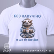 Футболка кавучіно злочіно, біла, складена на столі, фон світлий, принт з ведмедем-ленивцем у піжамі, який тримає чашку, текст зверху і знизу синього кольору, на комірці видна етикетка.