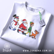 Футболка казковий настрій. Біла футболка, складена на поверхні, з принтом, що зображає Санту, сніговика, ельфа і оленя. На футболці видно етикетку з зеленим шнурком. Фон світлий, акцент на принті.