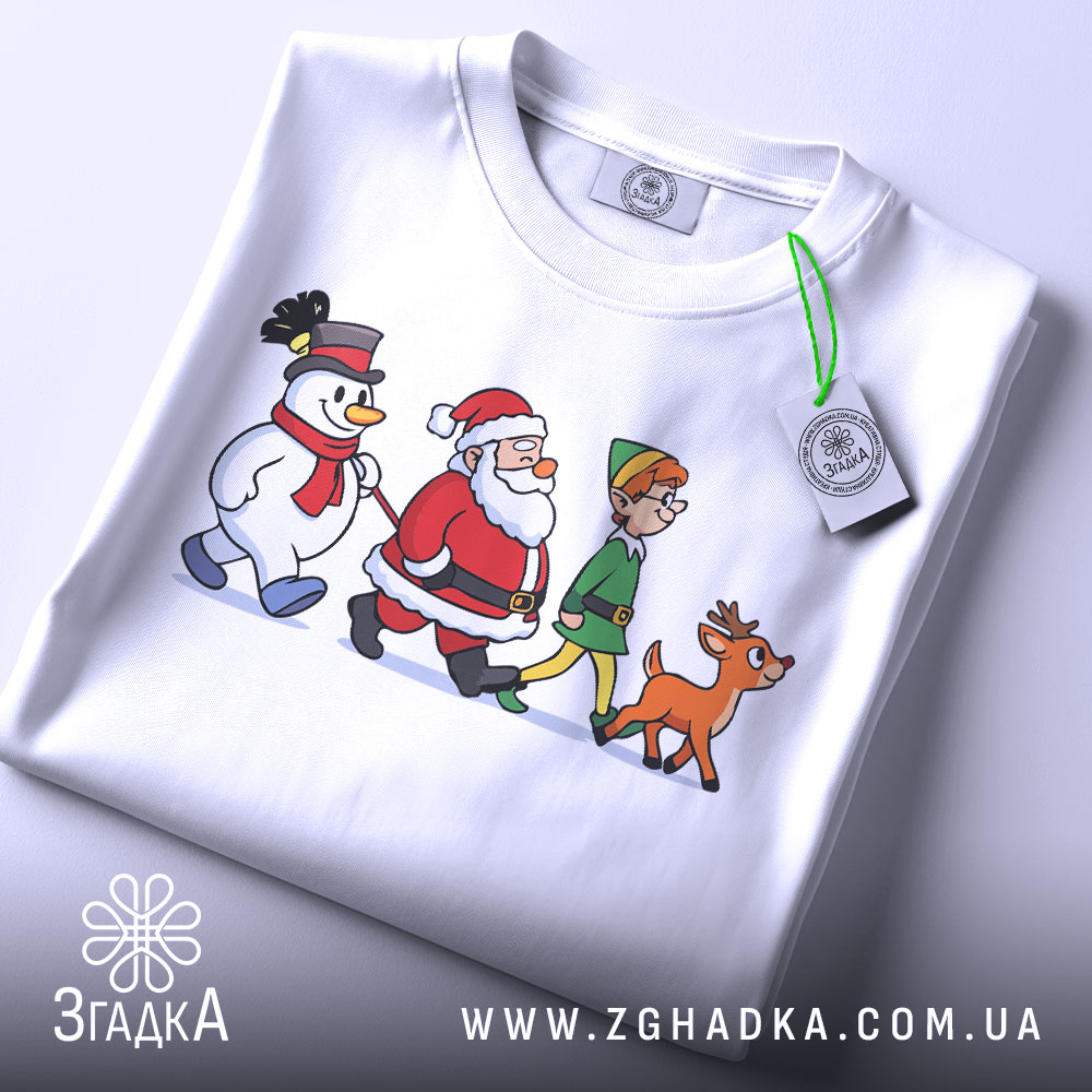 Футболка казковий настрій. Біла футболка, складена на поверхні, з принтом, що зображає Санту, сніговика, ельфа і оленя. На футболці видно етикетку з зеленим шнурком. Фон світлий, акцент на принті.