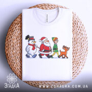 Футболка казковий настрій. Біла футболка, складена на плетеній основі, з принтом, що зображає Санту, сніговика, ельфа і оленя. Фон світлий, акцент на текстурі плетеного матеріалу.