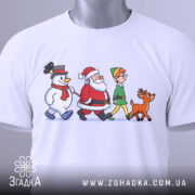 Футболка казковий настрій. Біла футболка, складена на поверхні, з принтом, що зображає Санту, сніговика, ельфа і оленя. На футболці видно етикетку з зеленим шнурком. Фон світлий, акцент на принті.