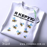 Футболка керую бджілочками, білого кольору, складена на поверхні, фон світло-сірий, принт з бджолами та написом "Я КЕРУЮ ЦИМИ БДЖІЛОЧКАМИ" в чорному кольорі, з зеленим ярликом.