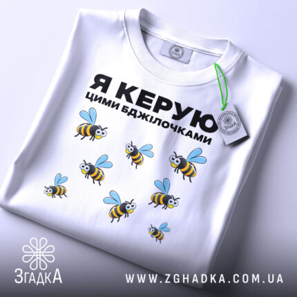 Футболка керую бджілочками, білого кольору, складена на поверхні, фон світло-сірий, принт з бджолами та написом "Я КЕРУЮ ЦИМИ БДЖІЛОЧКАМИ" в чорному кольорі, з зеленим ярликом.