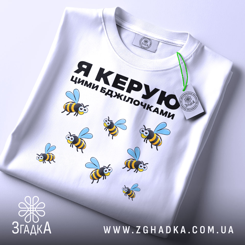 Футболка керую бджілочками, білого кольору, складена на поверхні, фон світло-сірий, принт з бджолами та написом "Я КЕРУЮ ЦИМИ БДЖІЛОЧКАМИ" в чорному кольорі, з зеленим ярликом.